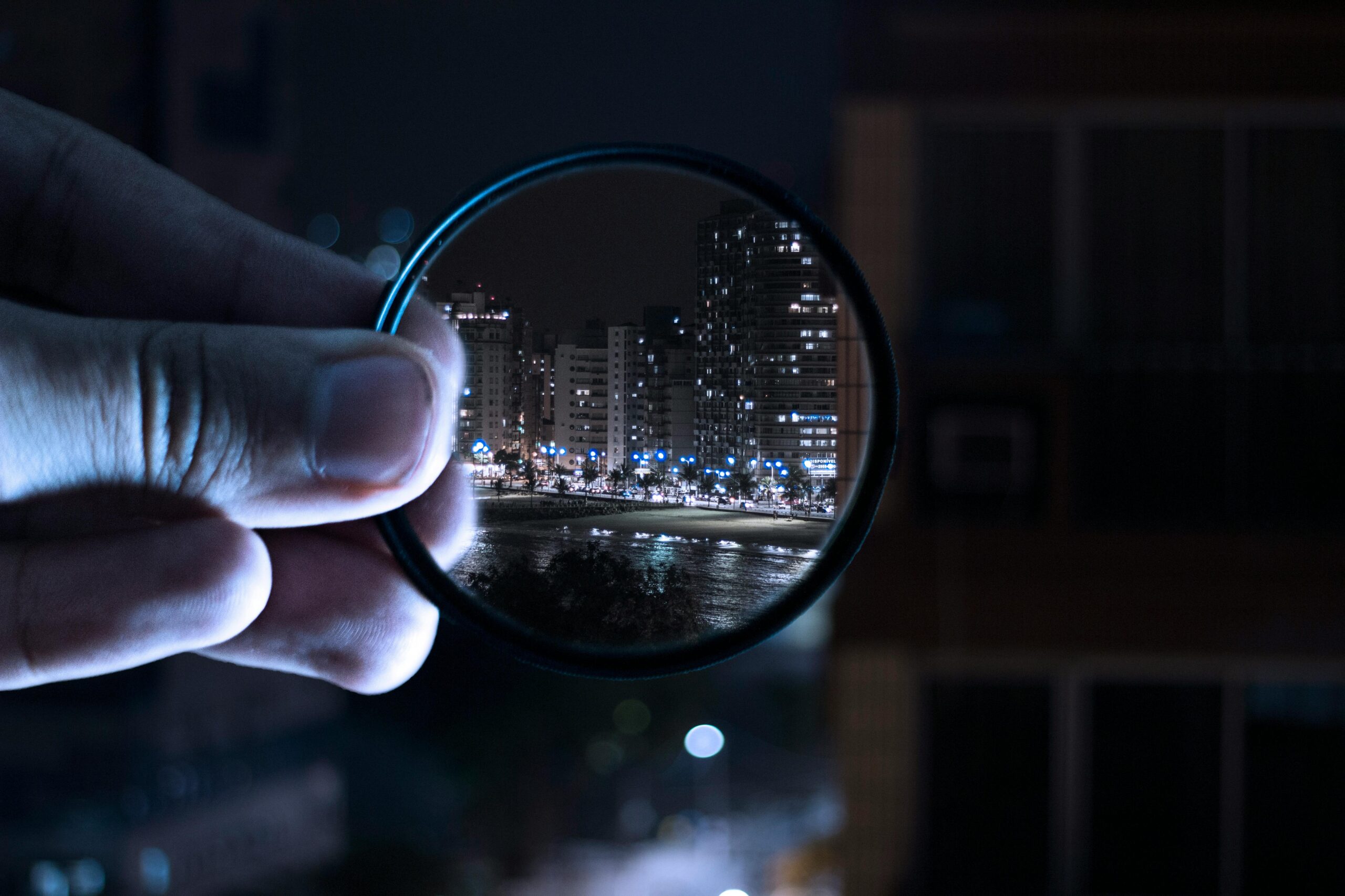 A round magnifying glass is held up by fingers from the left, with a city visible through the glass.