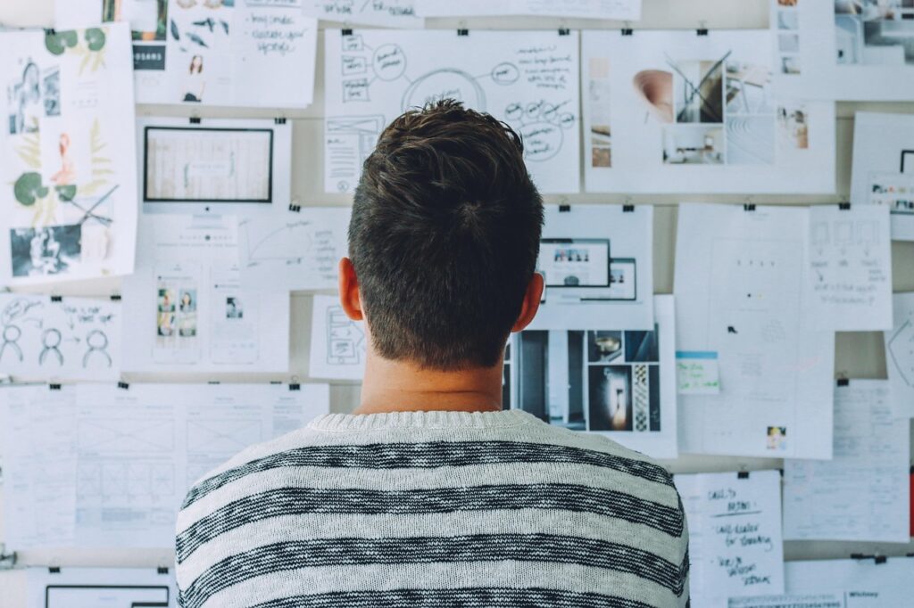 A white man with a striped sweater looks at a tack board filled with different documents