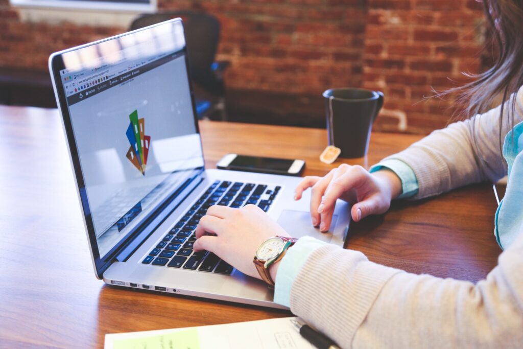A light skinned person types on a computer on a wooden table with a brick wall in the background.