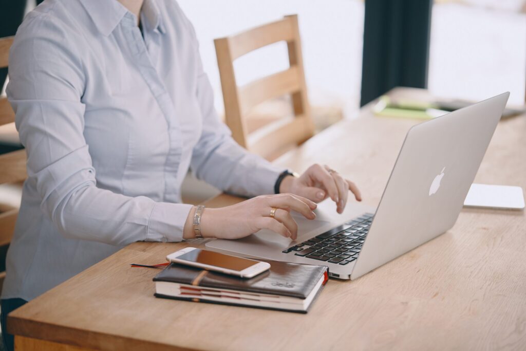 A light skinned person wearing a light blue button-up types at a laptop on a wooden table.