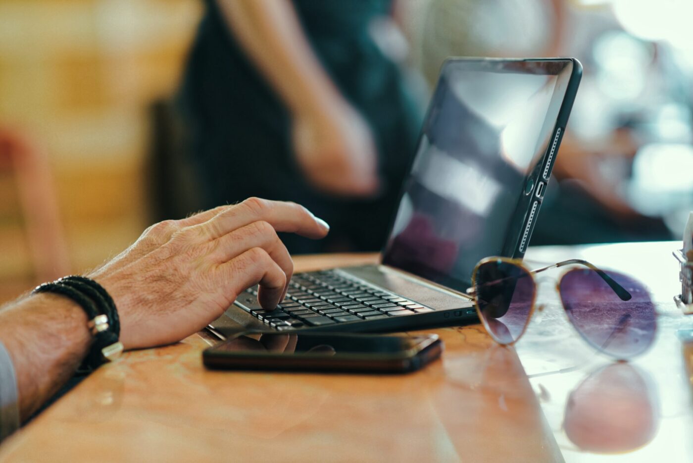 A light-skinned hand types on a keyboard, with sunglasses and a phone adjacent, on a wooden table.