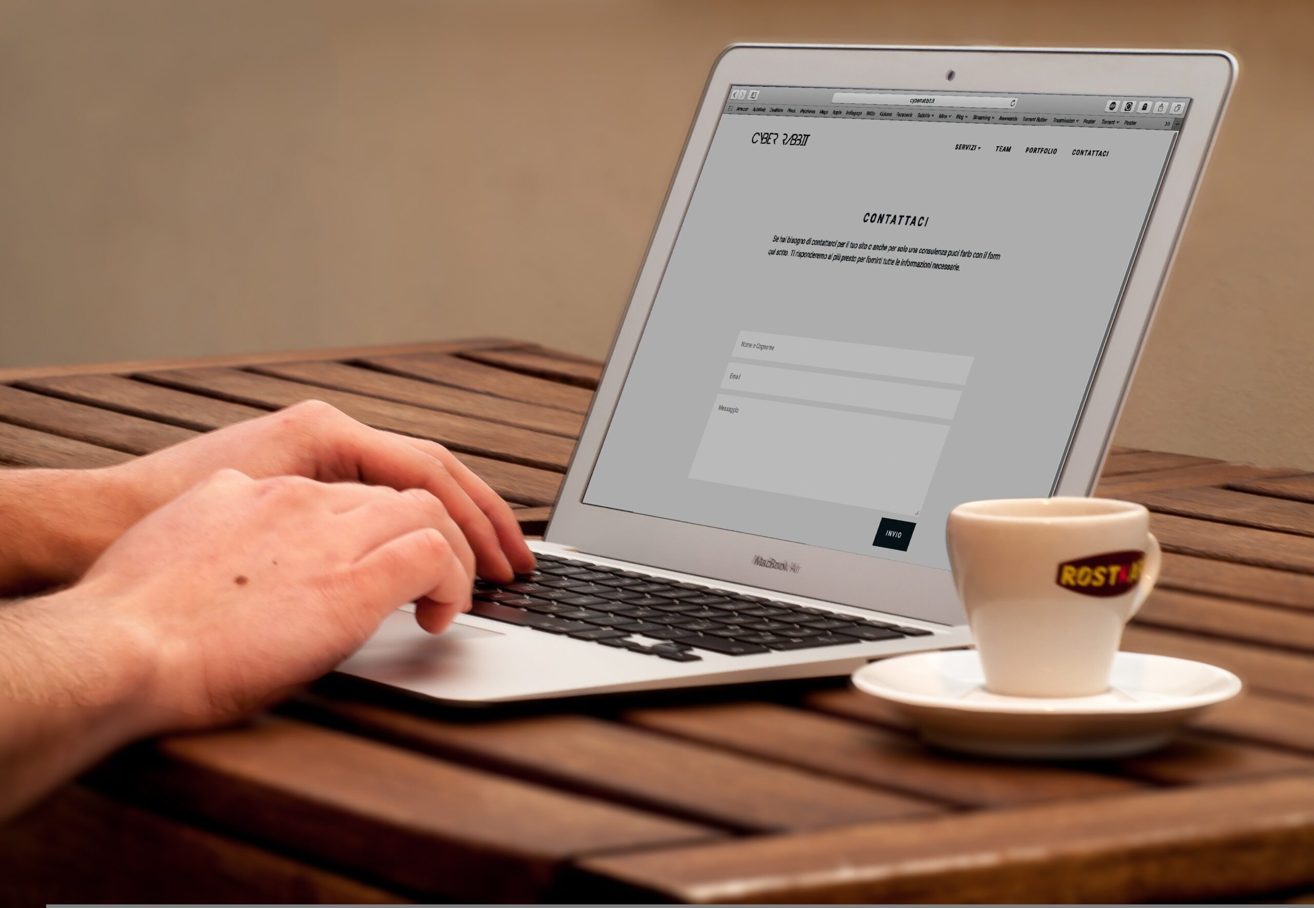 A pair of light-skinned hands types on a laptop on a wooden table, with a white coffee cup on a small plate nearby.