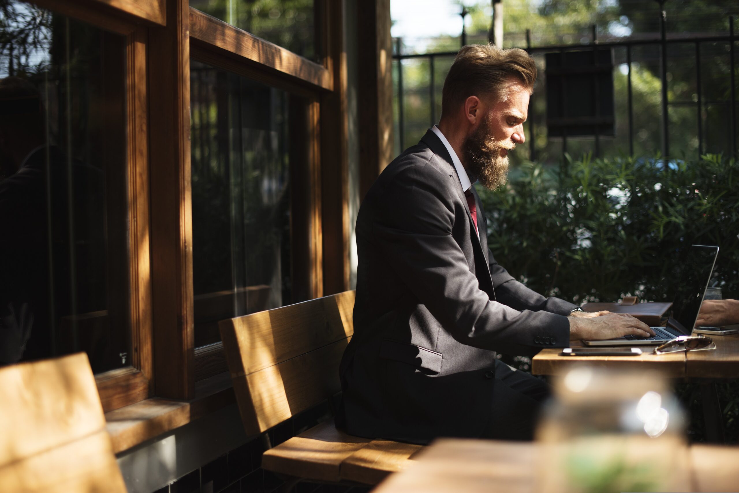 A bearded light-skinned man in a suit types on a laptop in a sunny outdoor seating area with plants in the background.