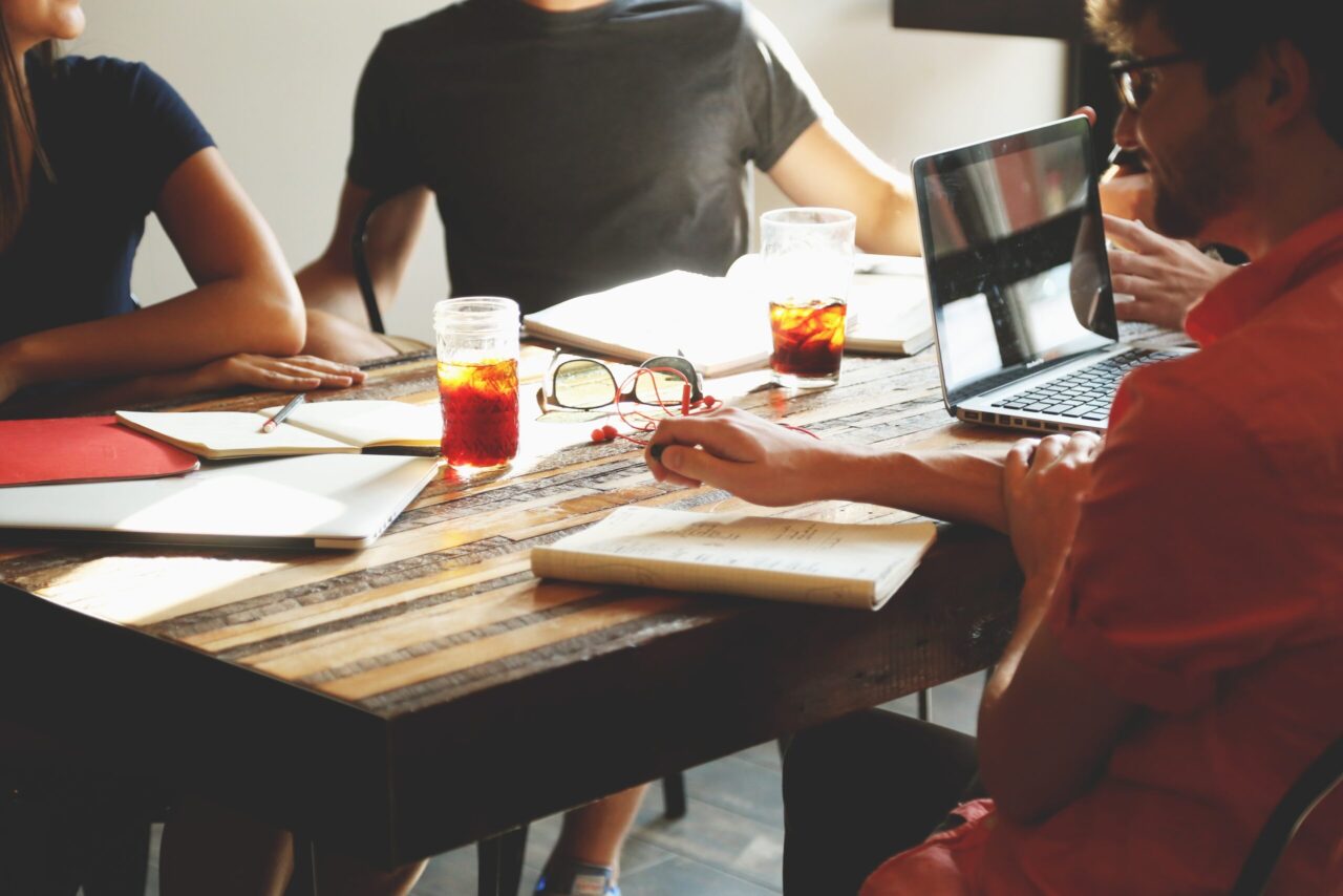 A group of people meet around a table, which is covered with a laptop, cups, and papers.