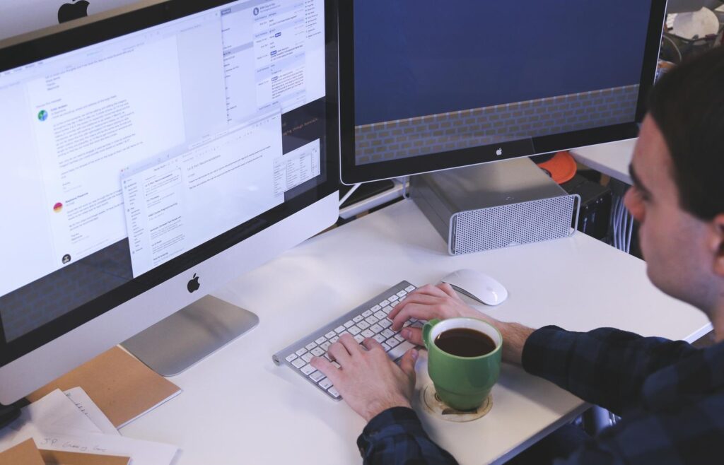 A light skinned man types on a keyboard in front of two monitors, with a coffee cup between his arms.