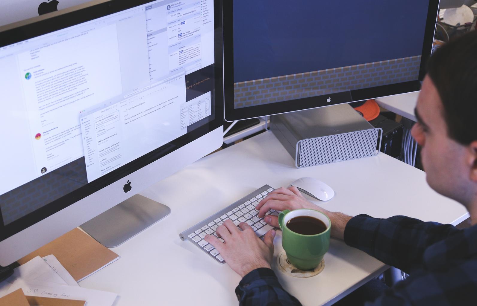 A light skinned man types on a keyboard in front of two monitors, with a coffee cup between his arms.